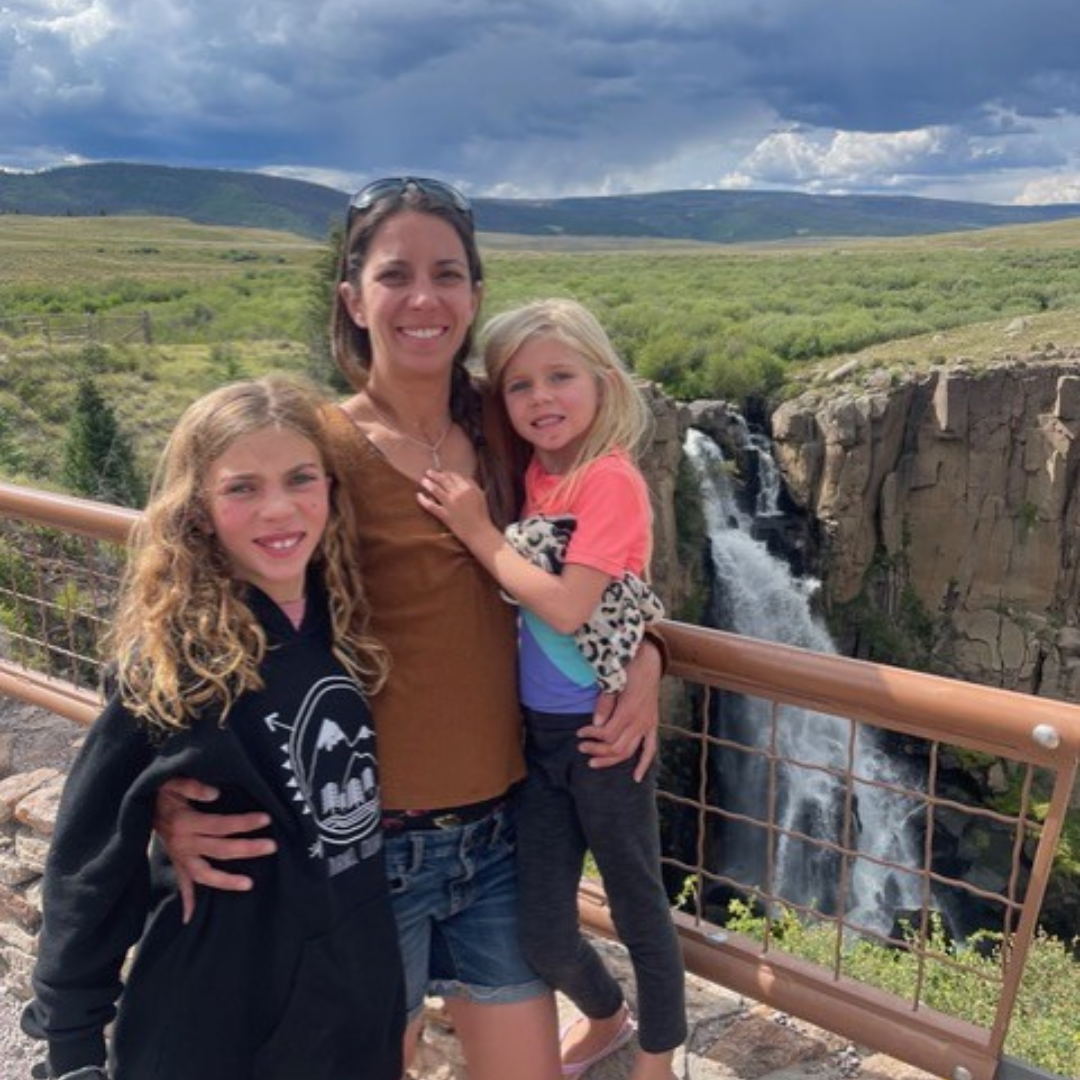 a mom and her two young girls pose for a photo in front of a slightly cloudy, outdoor mountain backdrop.