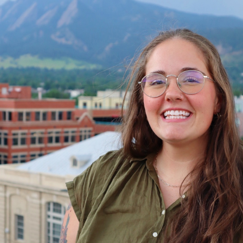 A smiling person with glasses standing in front of a mountain.
