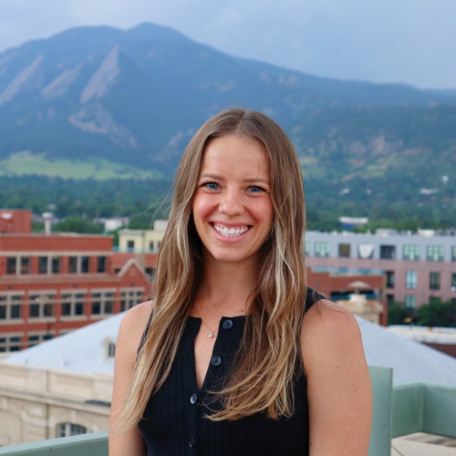 A smiling person standing on top of a building with mountains in the background.