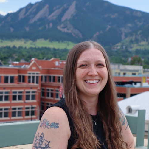 a person with long hair and tattoos sitting on a balcony with mountains in the background.