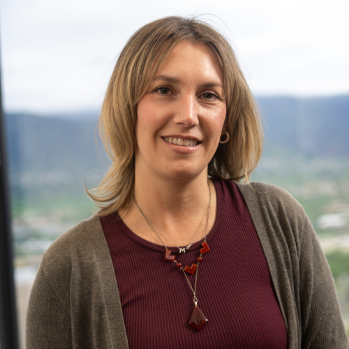 a person with a burgundy shirt and brown sweater standing in front of a window with mountains in the background.