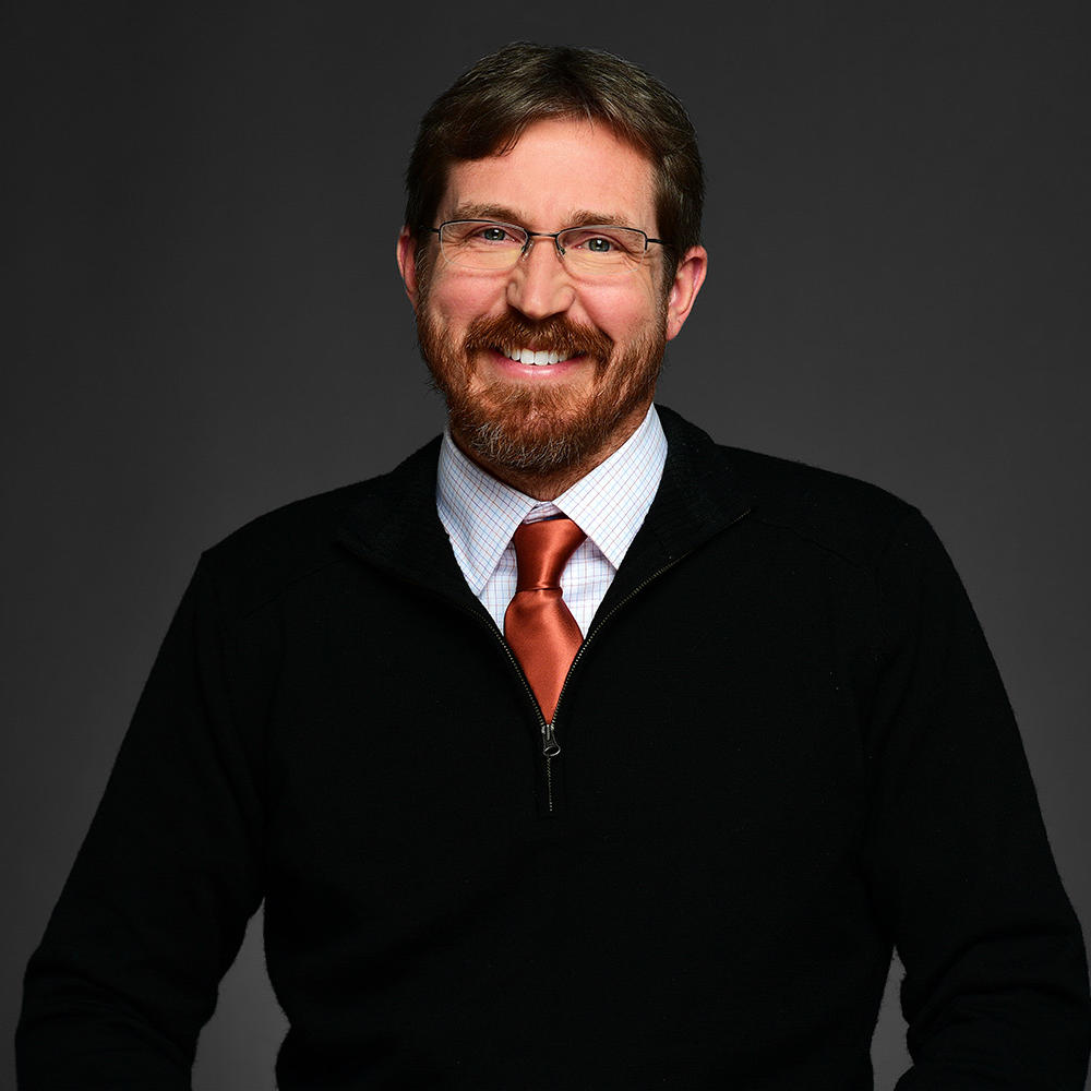 Eric Pynn is wearing a black jacket, white collard shirt, orange tie, and glasses. Eric has short hair and beard. The background is a dark gray.  