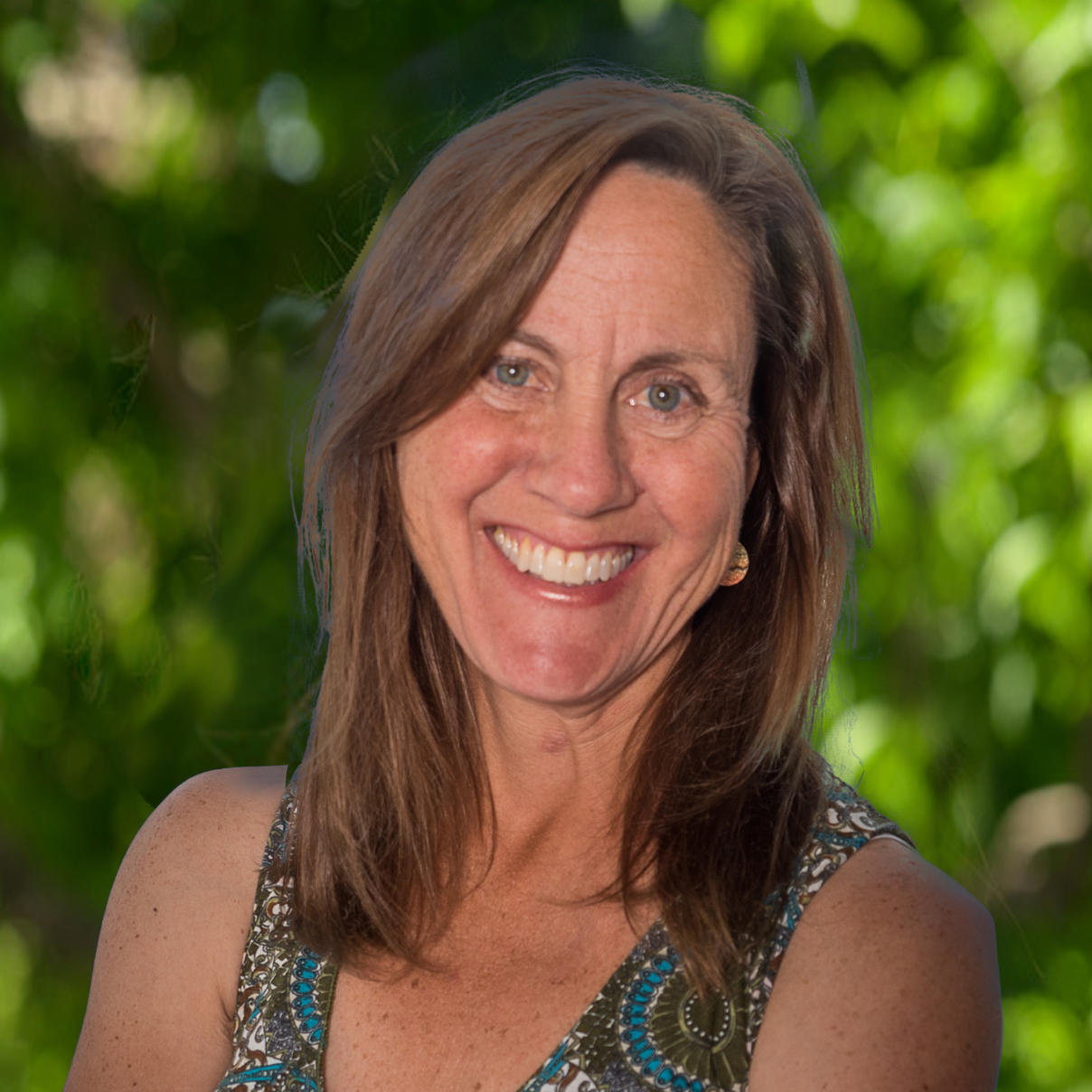 Angela has medium length light brown hair. She is wearing a gray and blue geometric pattern tank top. She is looking at the camera and has a green nature background. 