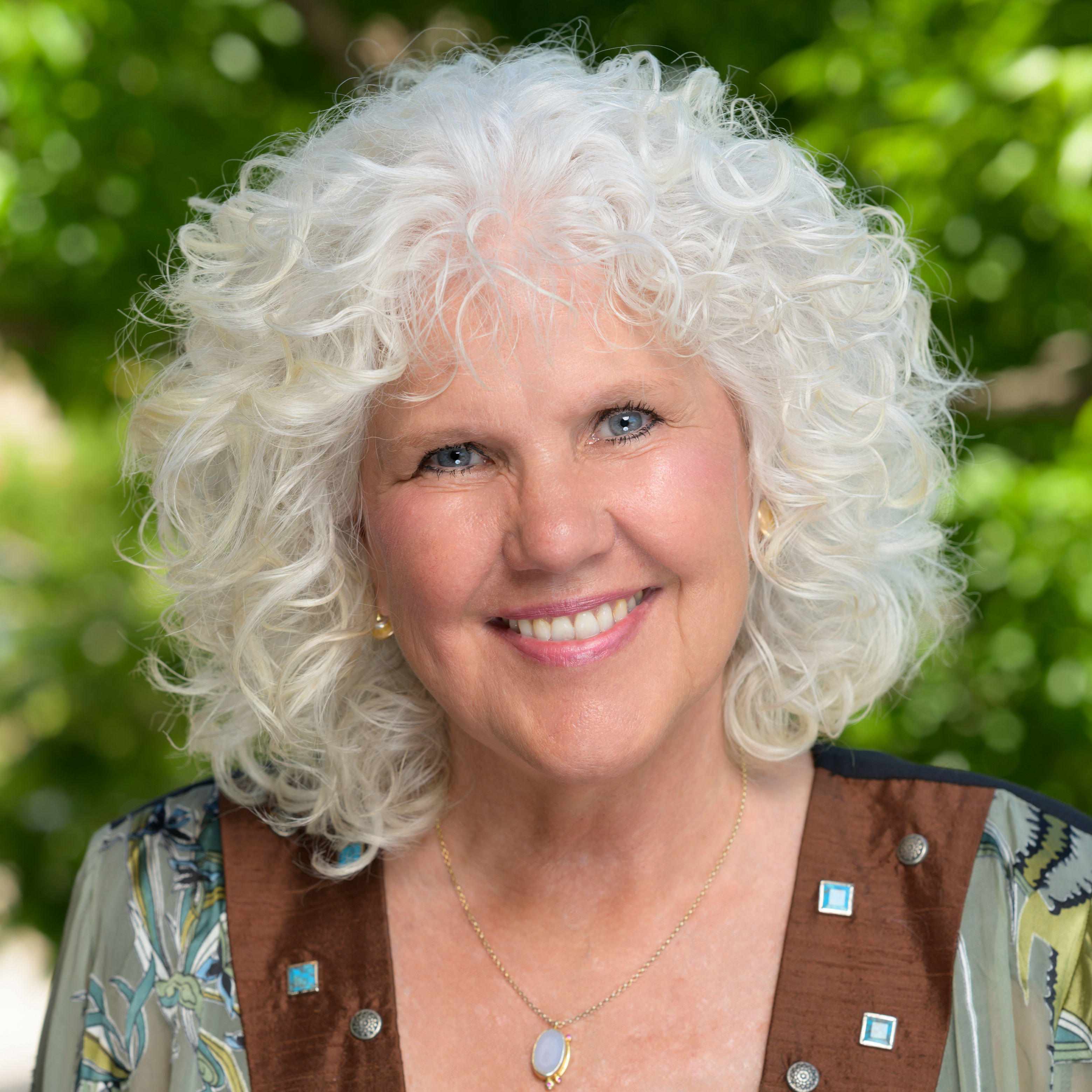 Jamie is looking at the camera and smiling. She has short-medium white curly hair that goes above her shoulders. She has a a a blue oval necklace, a blouse with floral patterns, the patterns include two brown strip with blue squares, and a black floral shirt underneath. Jamie has blue eyes and in standing in front of a green nature background.