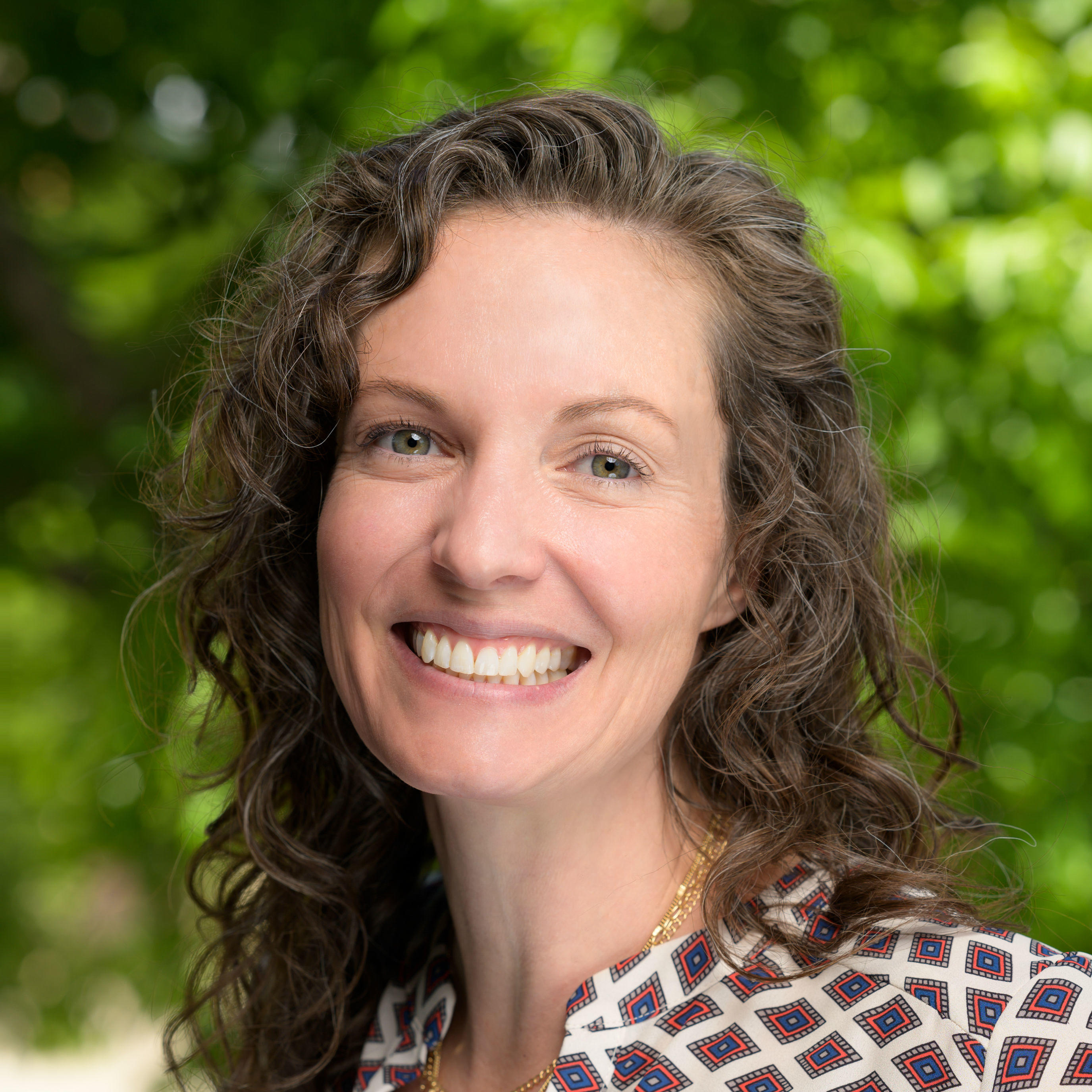 China is smiling and looking at the camera. She has long light brown curly hair and green eyes. She is wearing two gold necklaces with a circular pendant. She is also wearing a geometric diamond pattern white, grey, orange, and blue collard shirt. The background is blurred green nature with the corner left of the image having a blurred wall.