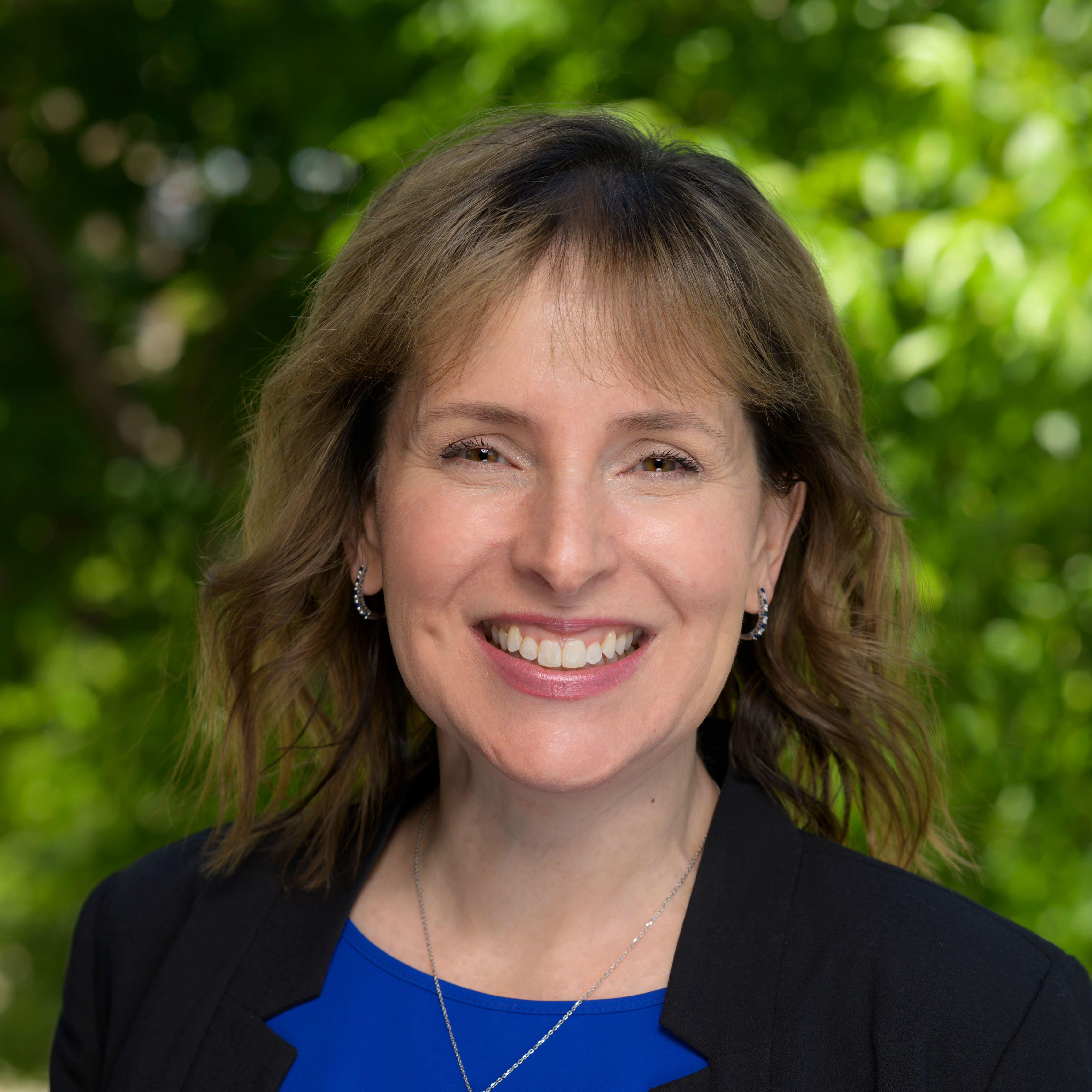 Hope has a light brown medium length hair. She is smiling and looking at the camera. She is wearing a black business jacket with a blue shirt underneath. She is also wearing silver with black hues earrings and necklace accessories. She has light hazel eyes and the background of the image is of green blurred nature. 