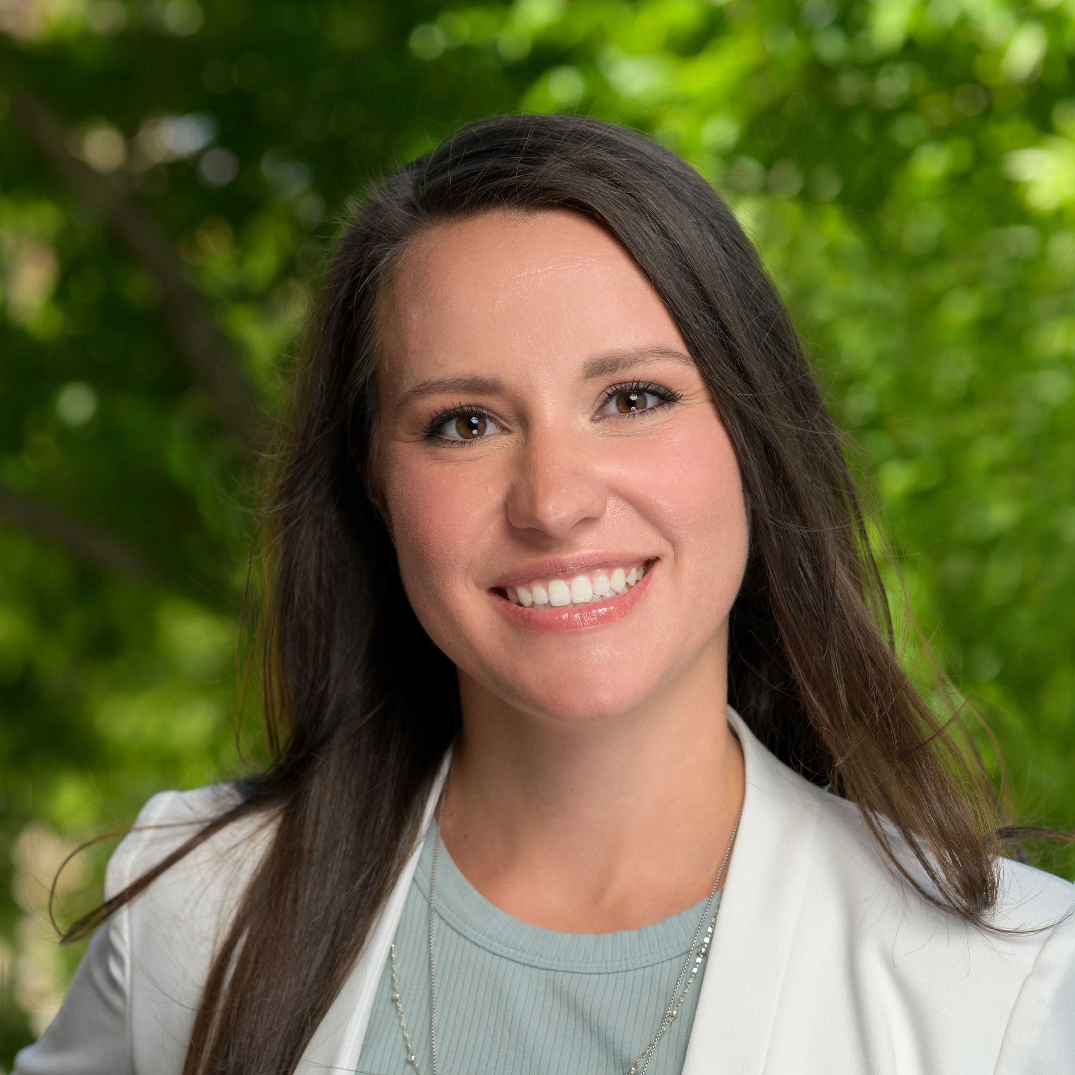 Lisa is smiling and looking at the camera. She has long brown hair and is wearing a white business jacket with a light green shirt underneath. She has light brown eyes and two silver necklaces with a turquoise pendant. The background of the image is blurred green nature. 