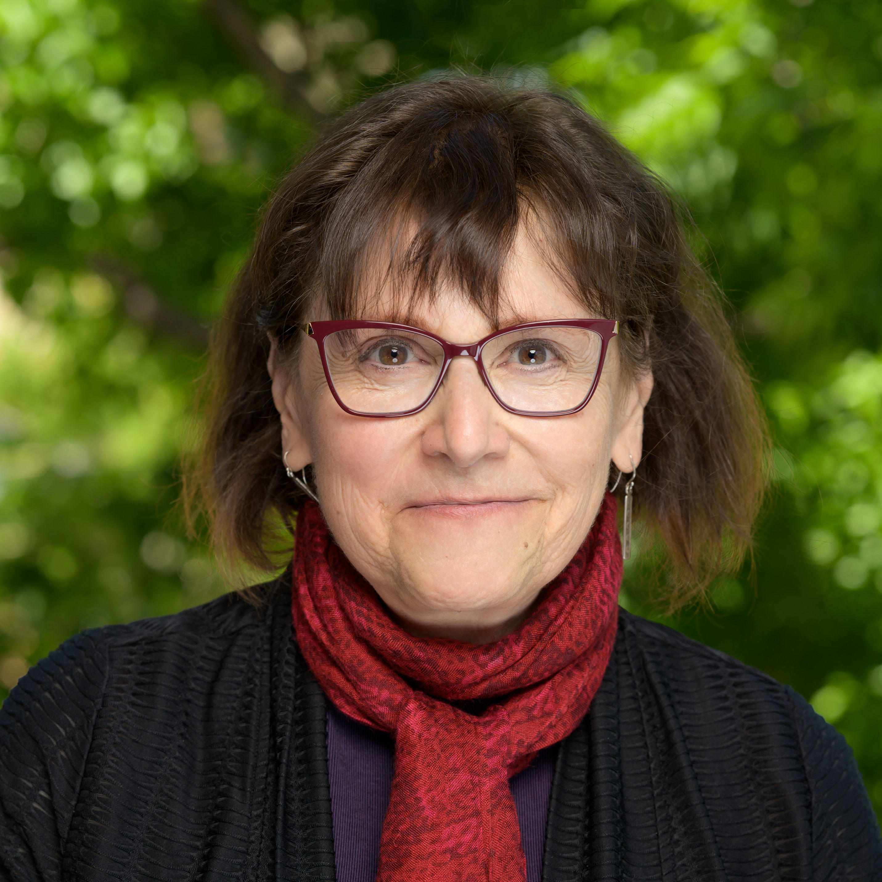 Sharon is smiling and looking at the camera. She has short brown hair with forehead bangs. She is wearing a red scarf,  burgundy glasses, vertical rectangular earrings, black cardigan, and purple shirt underneath. The background of the image is of blurred green nature. 