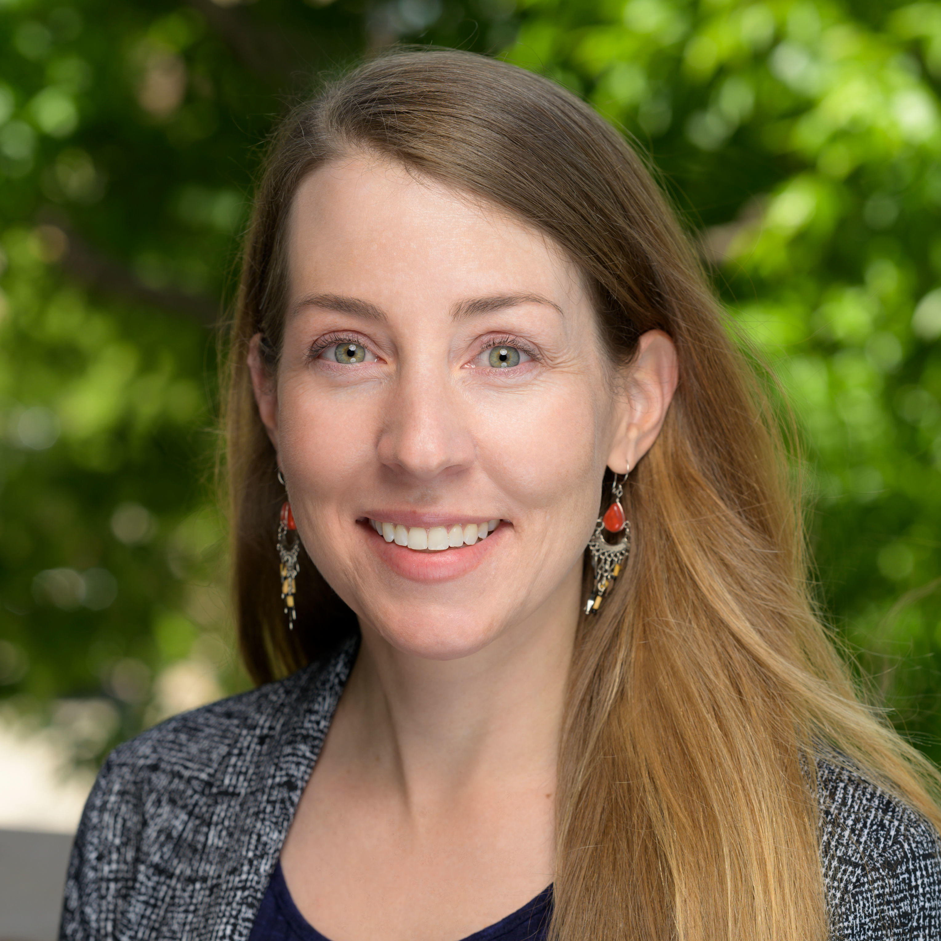 Erin has long dark blonde hair and green eyes. She is wearing chandelier earrings that are silver and orange-red. She is wearing a grey and black pattern business jacker with a dark blue vertical stripped shirt. The image background is of green blurred nature. 