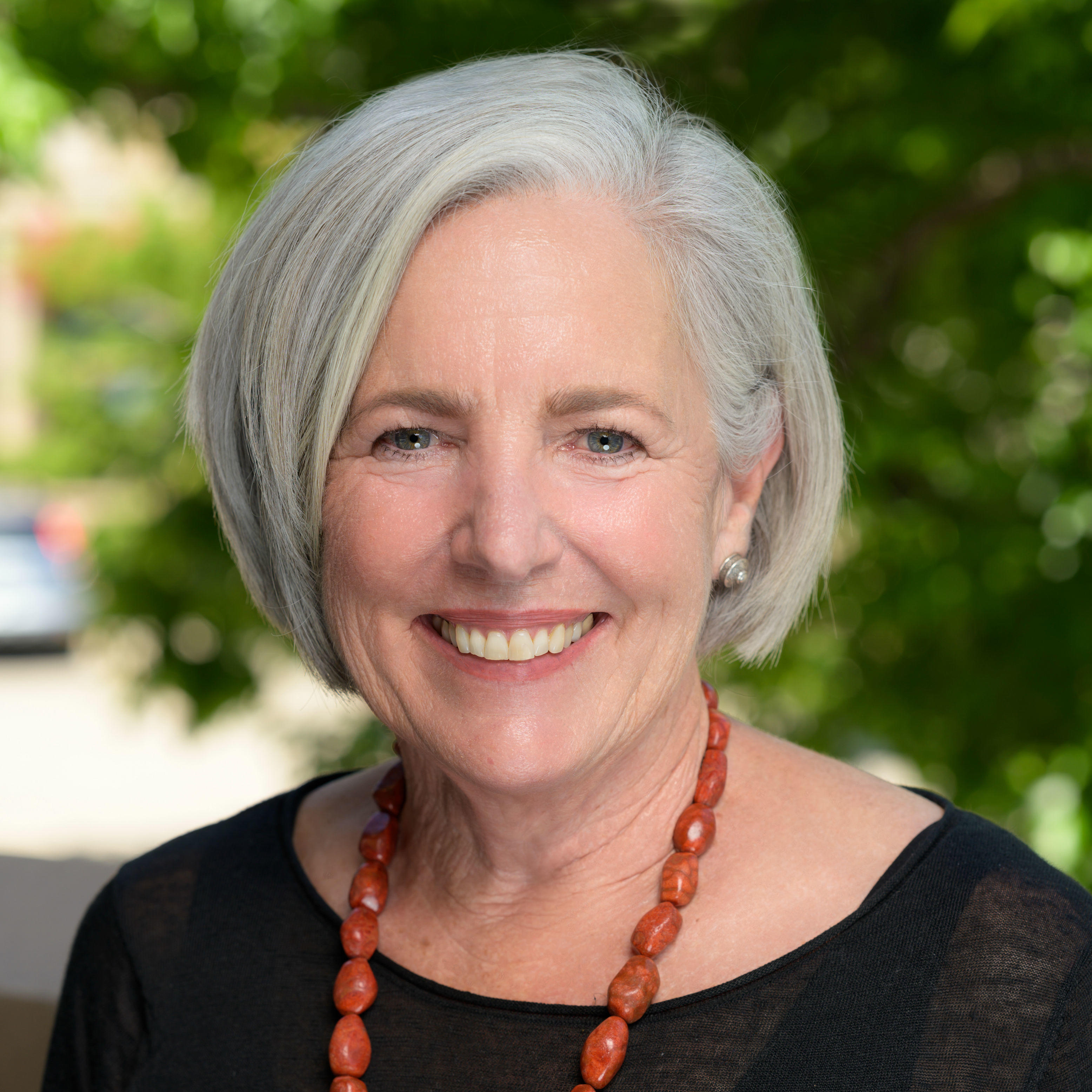 Nancy is smiling at the camera and has short length white hair. She has blue eyes and wearing circular silver earring. She is wearing a black blouse with a black tank top underneath and orange beaded necklace. The background is of blurred nature and on the left side is a blurred blue car. 