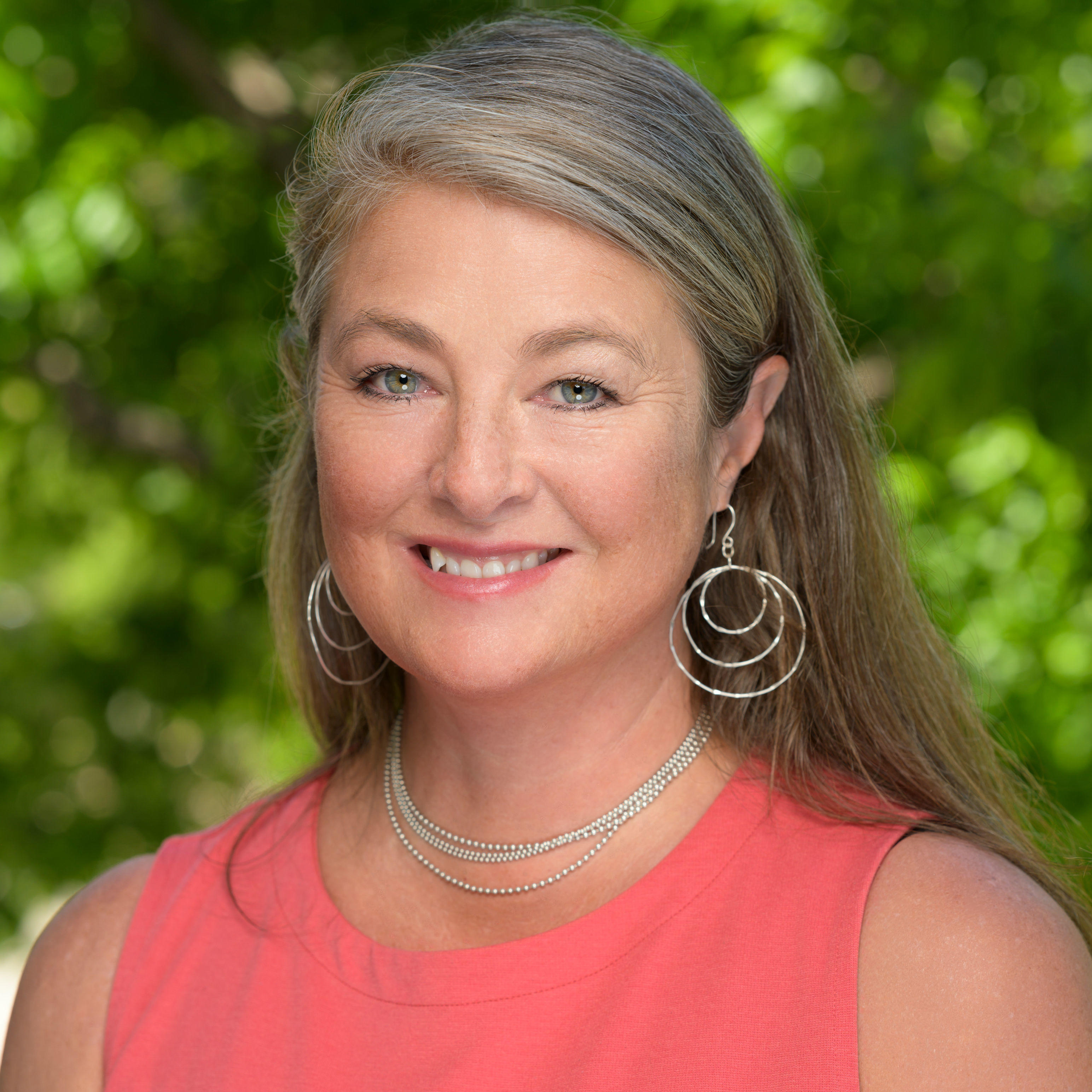 Mary is smiling and looking at the camera. She has long brown and silver hair. Mary has blue eyes, circular hoop earrings, silver necklace with multiple chains, and a pink tank top. The background of the image is blurred green nature. 