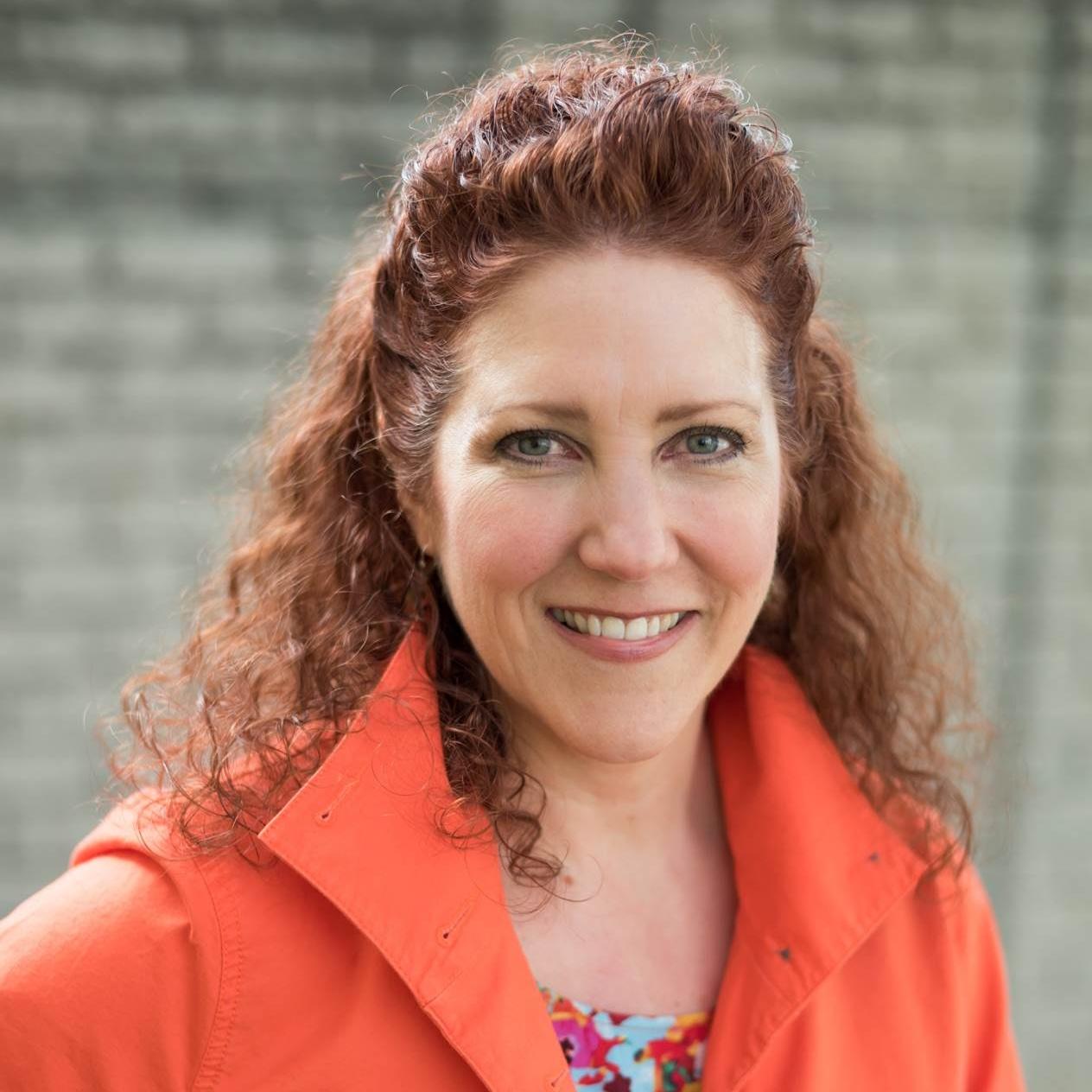 Tracy is smiling and looking at the camera. She has red, curly hair. She is wearing a flowery top with an orange coat over it against a blurred background of grey bricks.