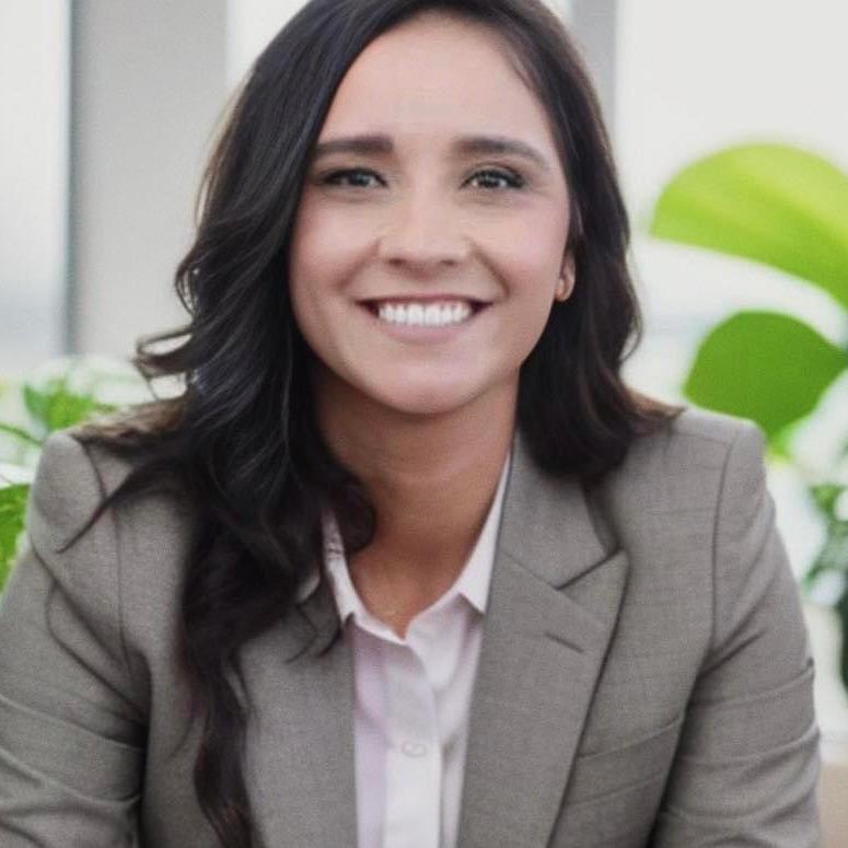 Sonia is smiling and looking at the camera. She has wavy brown hair and brown eyes. She is wearing a gray blazer and a white collared button shirt beneath with her sleeves pulled up. The background is a blurred office room with plants and bright daylight.
