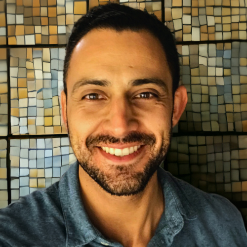 a smiling person wearing a blue shirt in front of a mosaic wall.