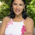 Andrea has medium short black hair and is wearing gold hoop earrings. She is smiling and has dark brown eyes. She has a necklace with the letter A on it and wearing a white tank top with roses on the tank top. The background is blurred trees. 