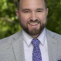 Brandon is smiling and looking at the camera. He has short brown hair with a short beard. He is wearing a grey business jacket, white collard shirt, a purple-blue floral pattern tie, and handkerchief in his business jacket chest pocket of the same pattern of his tie. Brandon's background image is of blurred green nature. 