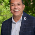 Brian is smiling and looking at the camera. He had short black and grey hair. The top of his hair is combed back and he is wearing a square pattern navy business jacket with a circular pin that says, "Shop Small Pueblo, Colorado." He also has a white buttoned collard shirt. The background is of blurred green tree. 