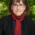Sharon is smiling and looking at the camera. She has short brown hair with forehead bangs. She is wearing a red scarf,  burgundy glasses, vertical rectangular earrings, black cardigan, and purple shirt underneath. The background of the image is of blurred green nature. 