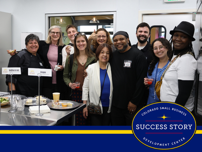 A group of people pose for a photo in what appears to be a catering or food service setting. Several individuals are smiling and holding drinks, with some food items visible on a counter in the foreground.