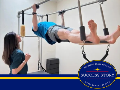A person in a blue shirt and gray shorts performs a Pilates exercise on a reformer apparatus, hanging from a horizontal bar with their feet supported on a wooden board with straps.