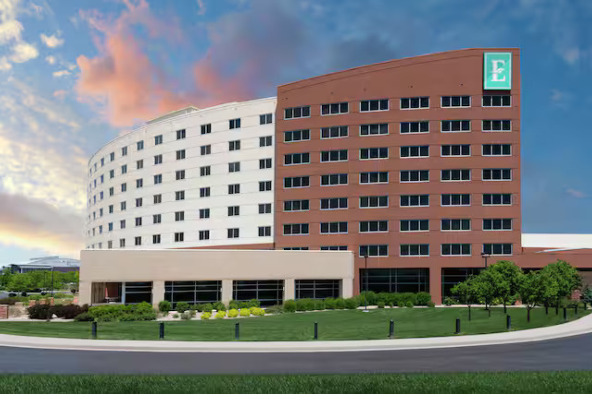 A modern hotel building with a curved white section and a rectangular brick section, featuring numerous windows and a green "E" logo on the brick facade. The building is surrounded by a manicured lawn and trees under a dramatic sky with pink and orange clouds.