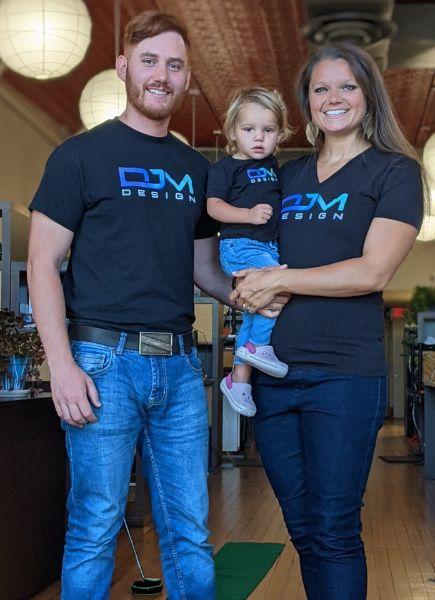 Image of Daniël-James van den Berg and his family. He is the owner of DJM Design. He is standing with a woman next to him. The woman is holding a small child in between them. They are smiling and in an office setting. They are wearing jeans and black shirts with the DJM Design logo. The logo is bold in blue and slim font for design underneath. 