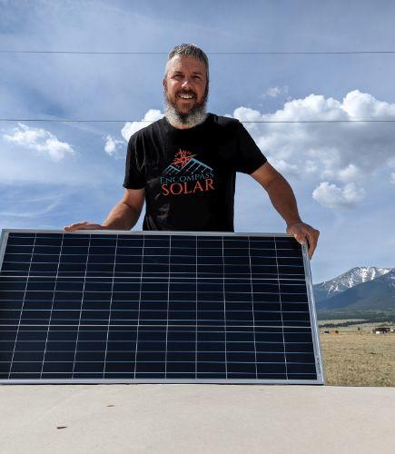 Image of Kenneth Tapp, owner of Encompass Solar. He is outside with a background of the mountains. He is holding a square solar panel and is wearing a black shirt with company logo. He has gray hair and is smiling at the camera. 