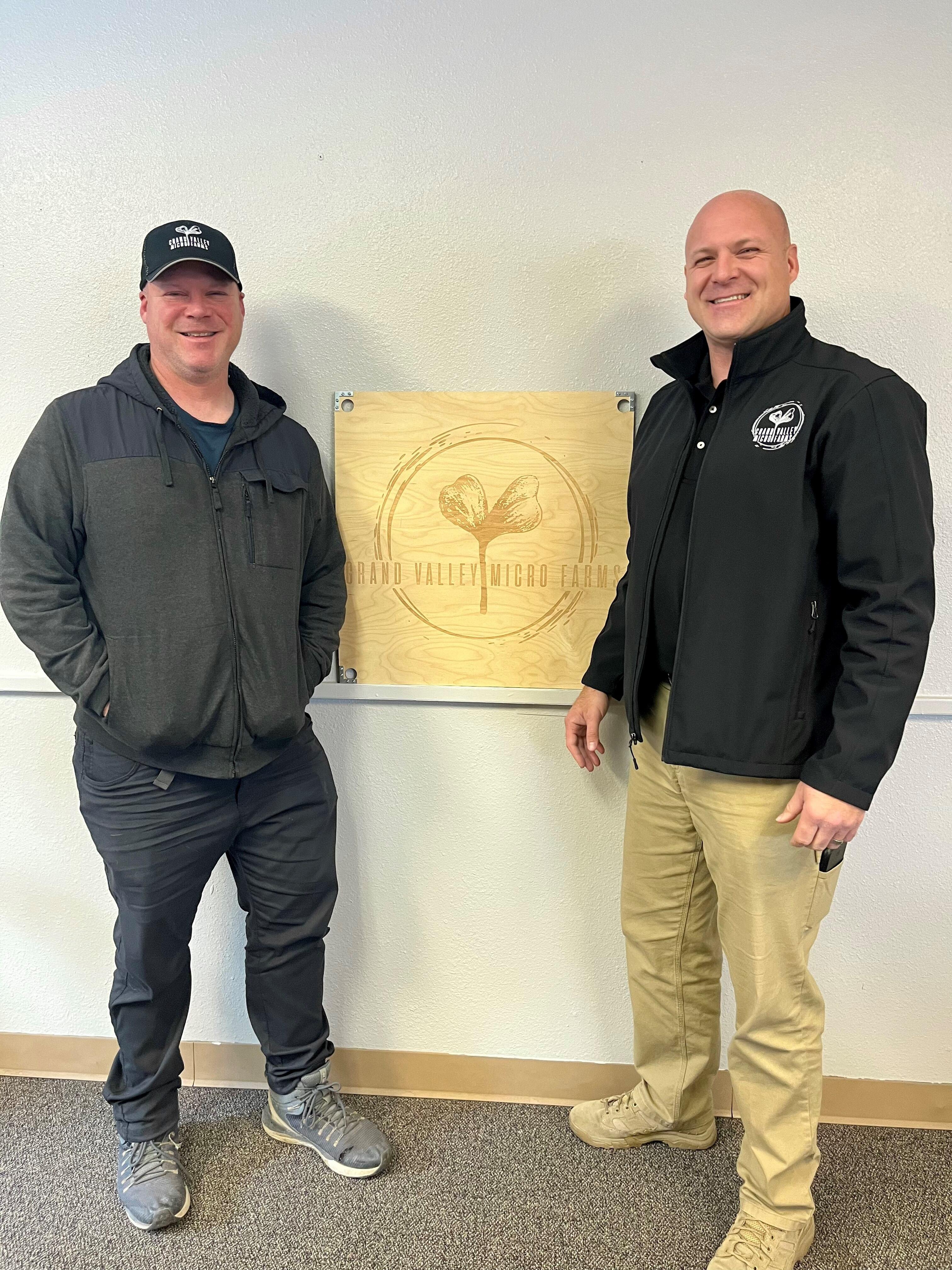 Image of Nate and Phil Strouse. They are brothers who are co-owners of Grand Valley Micro Farms. They are standing in the photo and in the middle is a wooden square of their logo. The logo is a circle with their company name in the middle and a plant growing in the middle. 