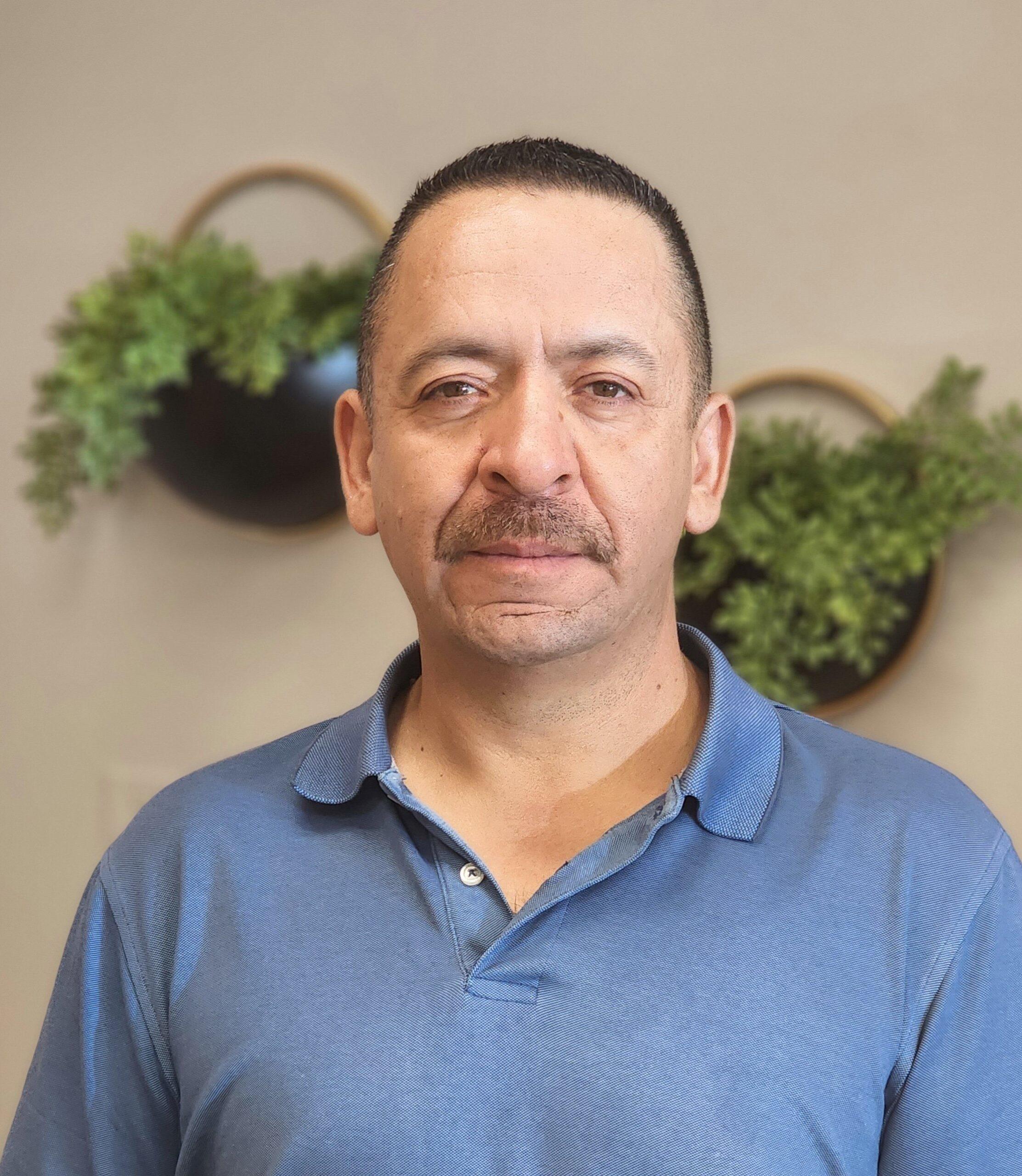 Head shot Image of Jorge Torres. He has a buzzcut hair style. He's got a shaven mustache and wearing a blue collard shirt. There background is a beige wall with two plants hanging on the wall. 
