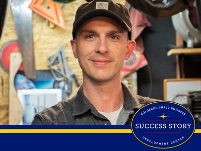 A person wearing a baseball cap and a collared shirt smiles at the camera in a workshop with tools in the background.