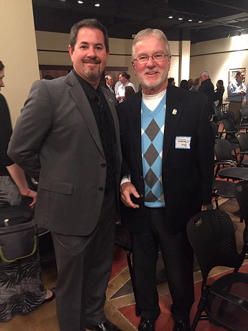 Image of Mike Tipton and Ron OHerron. They are both standing next to each other. The man on the left is slightly taller than the man on the right. The man on the left is wearing a gray suit and the man on the right has a black suit with blue diamond shirt underneath. The man on the right has glasses. The setting is of a  conference event with people standing and talking in the background. 