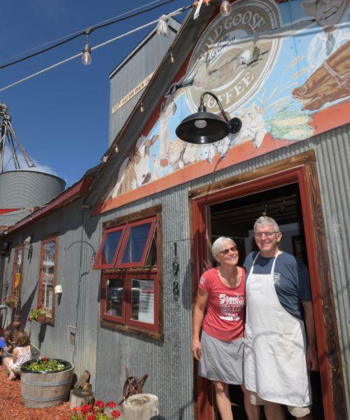 Tammie and Patrick Delaney are standing together in front of a door. The background has the Hayden Granary. There are children playing on the side of the building with adults sitting at a table.