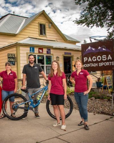 Image of Pagosa Mountain Sports Owners. The image has a yellow house as the background in an outdoor setting. The yello house is the business location of Pagosa Mountain Sports and the four owners are in the front. There is a gazebo on the side of the house. The owners are Jake and Janine Emmets, Amanda Gadomski, and Coquette Collins. The four women are wearing a burgandy polo shirt and the man is wearing a gray polo shirt. The man has a mountain blue bike and the woman on the left is holding a tube float  