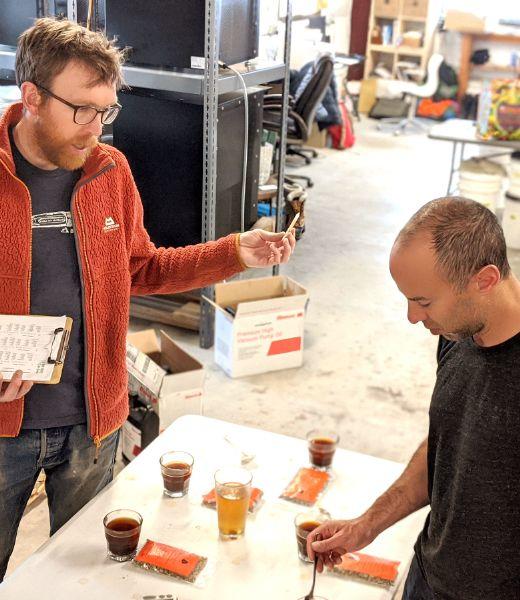 Image of Sam Higby and Mark Drucker. They are discussing around a table with five cups. Sam has glasses, an orange jacket, jeans, a clipboard, and a beard. Mark is wearing a black shirt and looking at the cup. The background is a  a manufacturing setting.