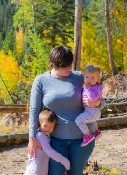 Image of Shelby Brandes, owner of High Plains Dice and Games. She is in an outdoor setting with two children. One of the children is hugging her leg and the other is a baby that she is holding. Shelby is looking at the baby and hugging the child below. There is a tree that fell behind them with green and yellow trees. 