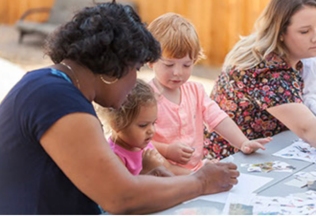 Female teacher in a blue dress helping pre-school-aged children with an activity.