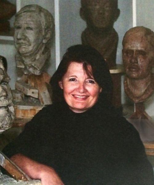 Image of Doctor Theresa Ferg. She has black hair and a black sweater. She is smiling and looking at the camera with her left arm rested on the edge of a table. Next to her there are head like sculpture models. There are six heads behind her with gray and brown colors. 