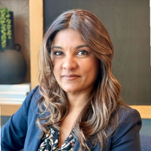 A person with long brown hair and a navy blazer over a floral top looks directly at the camera.