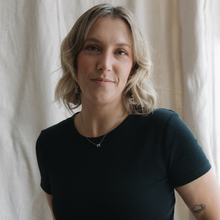 A person with blonde, wavy hair wearing a dark t-shirt and a delicate necklace with a small pendant.
