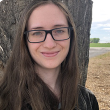 A person with glasses and long brown hair smiles in front of a tree.