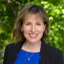 A smiling person with shoulder-length hair, wearing a blue top and black blazer, is set against a backdrop of green foliage.