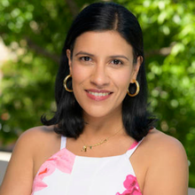 A smiling individual with dark hair and gold hoop earrings wears a floral top and a gold necklace.
