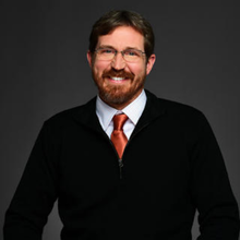 A smiling person with glasses and a beard, wearing a black sweater and an orange tie, poses against a gray background.