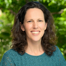 A person with shoulder-length wavy hair smiles in front of a blurry green background.