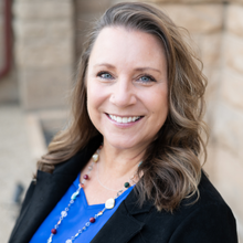 A person with blue eyes smiles, wearing a blue top, a black blazer, and a colorful beaded necklace.