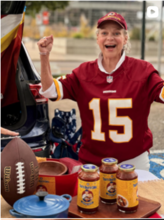 Tad Tandler is proudly cheering in a football jersey that is red, white and yellow with "15" on the front. In front of her there is a table with 3 jars of Tad's Dad's Chili, a blue crockpot, and a football.