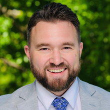 A smiling person with a beard wearing a grey jacket and floral patterned tie.