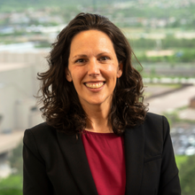 A person with shoulder-length dark hair smiles in front of a window, wearing a black blazer and a maroon shirt.