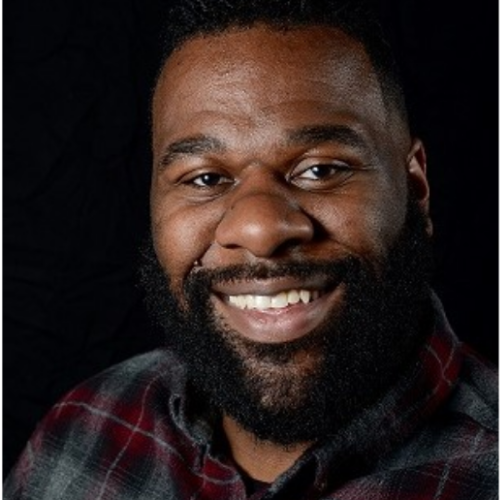 A close-up portrait of a person with a beard, wearing a plaid shirt against a dark background.